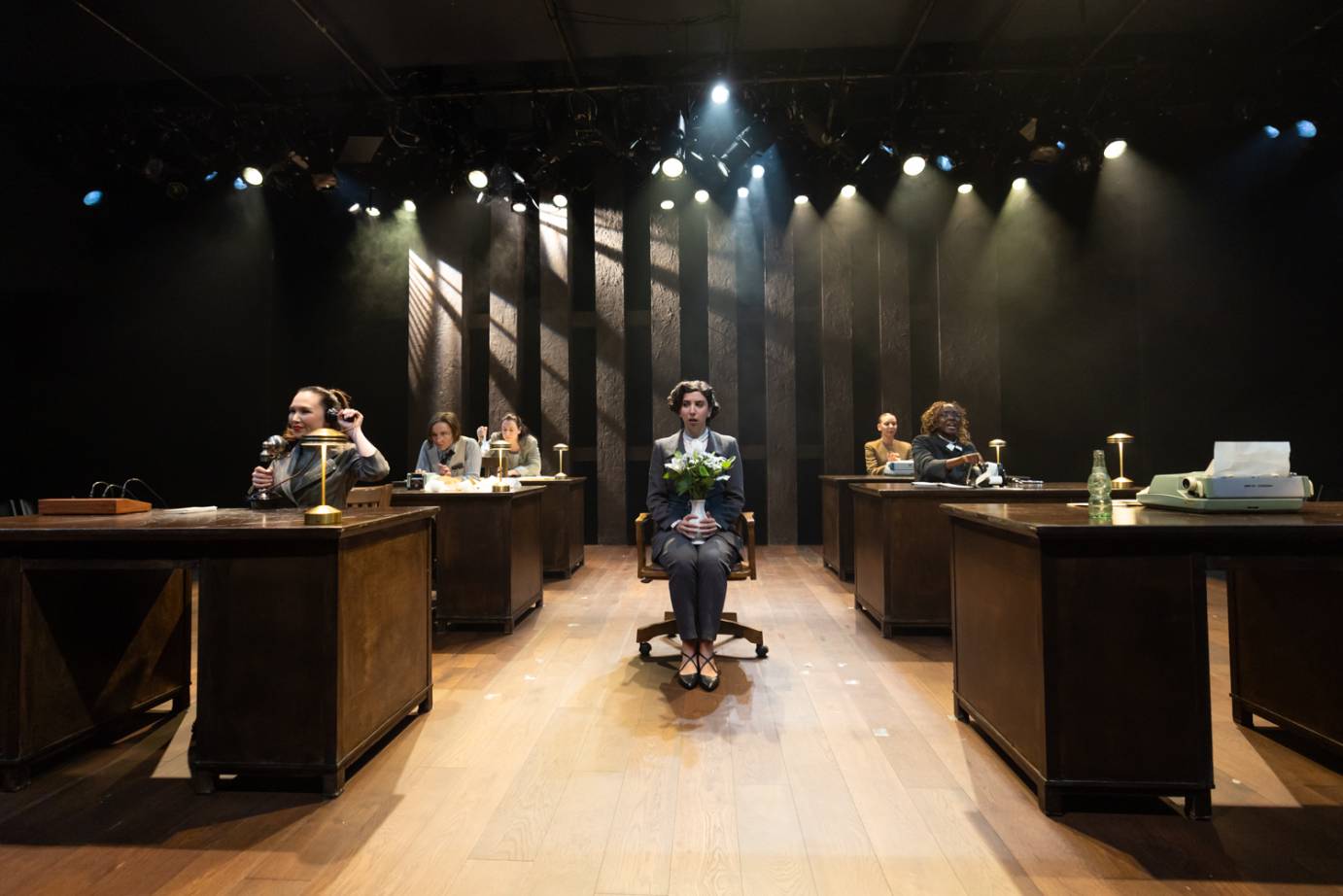 A seated woman wearing a grey suit with a bouquet of flowers in her lap, her face one of consternation and confusion, is seated between desks where workers sit accomplishing tasks.