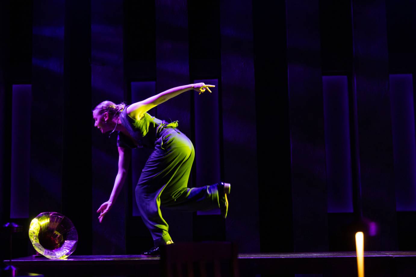 A blonde-haired woman on top of a desk, lit in green, bends forward with arms reaching on the diagonal and feet in a tapping run.