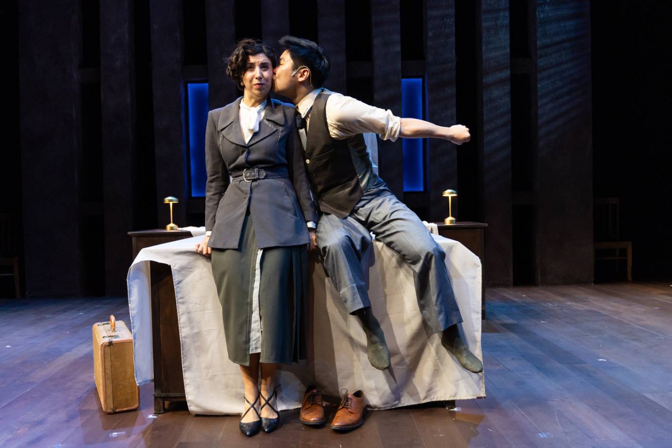 The protagonist woman in grey suit is kissed by a man in vest and white shirt as they are seated on a bed. Her face looks dismayed.