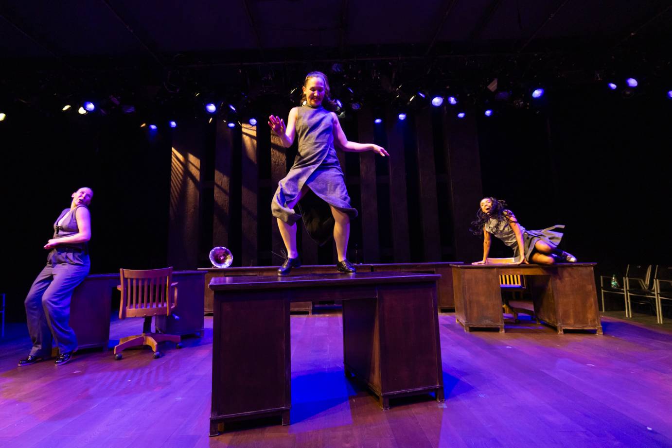 A woman exuberantly smiles as she taps on top of a desk, legs slightly apart and elbows to her waist. Two performers are on either side in the background, one atop a desk in a side crouch and the other standing with back to desk. Both look up at the central dancer.