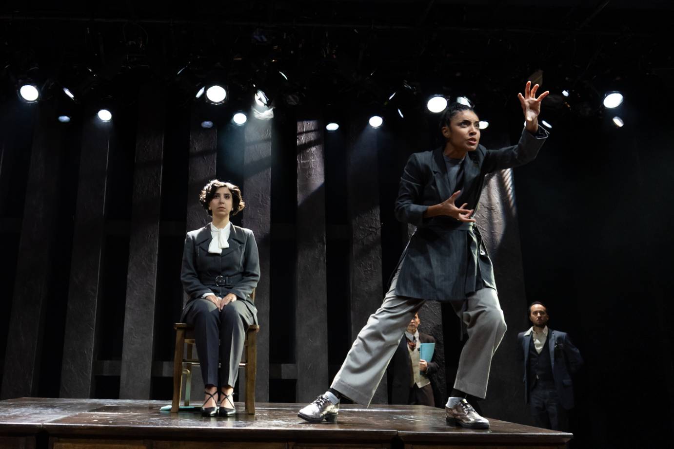 The protagonist in grey suit sits stoically facing forward on a chair while another woman looks to the diagonal, leaning forward, hands raised fingers splayed.