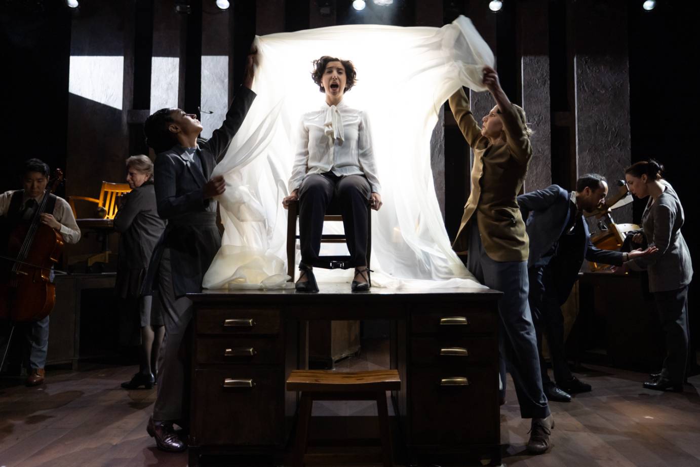 The seated protagonist in white bowed shirt and grey slacks, mouth open, is being shrouded by two performers with a swath of white fabric.