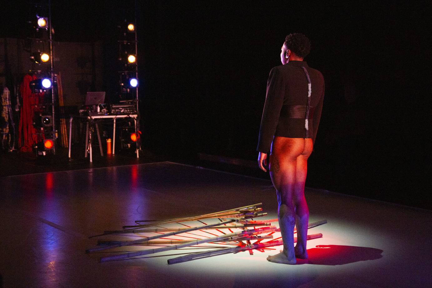 A man, bare-bottomed and wearing a black jacket, faces toward a stick sculpture