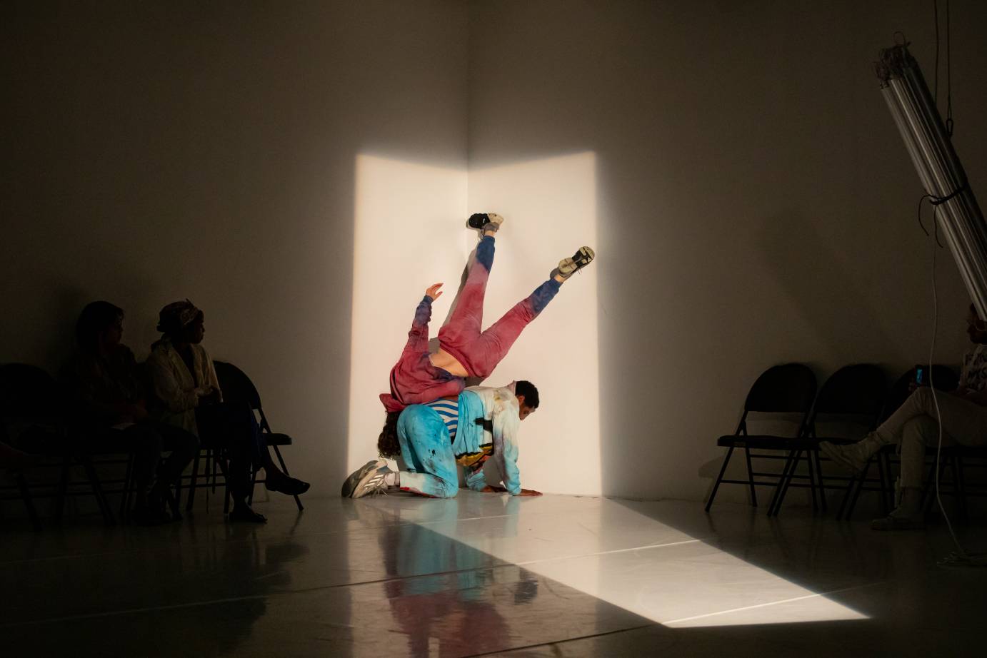 A woman in pink sweats upside down perched on the back of a kneeling man in torquoise sweats framed in a rectangle of light against a white wall.