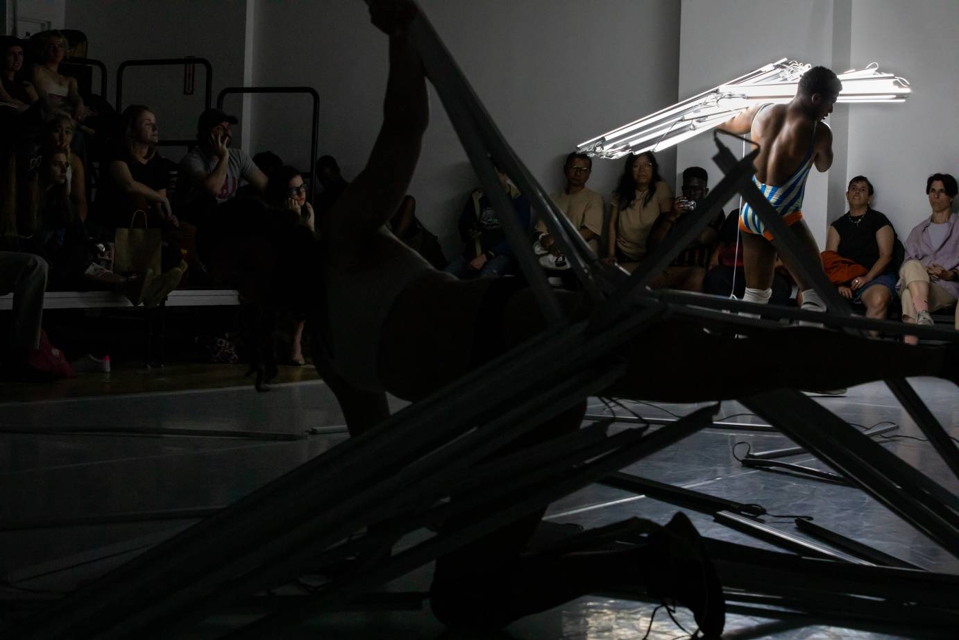 In a dimly lit space, a bare-backed man carries a lit bundle of slats over his shoulder amidst a scene of destruction. A barely lit figure on the floor holds up part of the set.