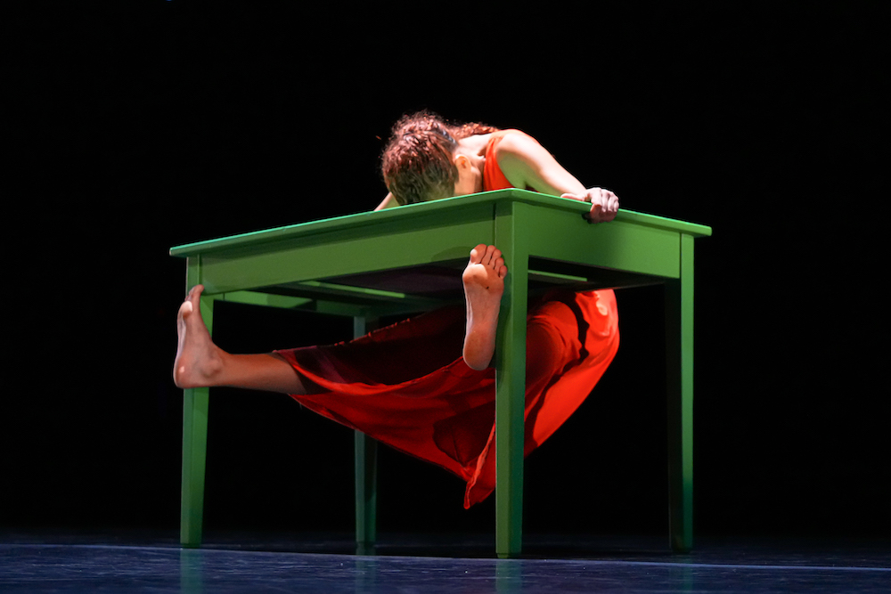 light skinned woman in bright orange slip dress leans her torso across a green table while sticking her barefeet out from under it 