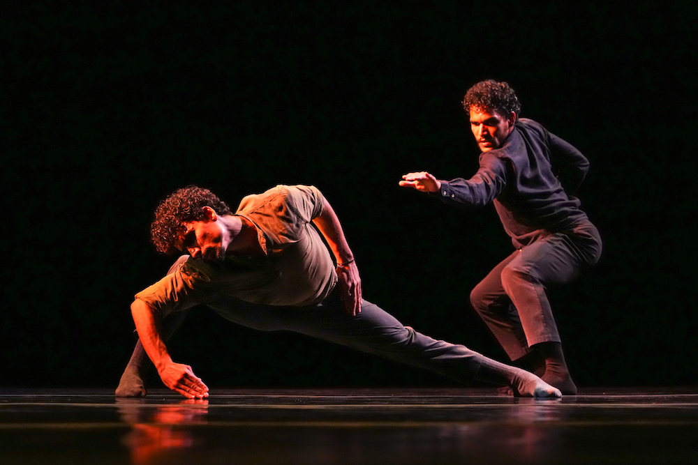 one man in a low right leaning lunge stares at the stage floor as his hand grazes it... another man behind him, all in black, crouches low and reaches his left arm out to the pensive man on stage  