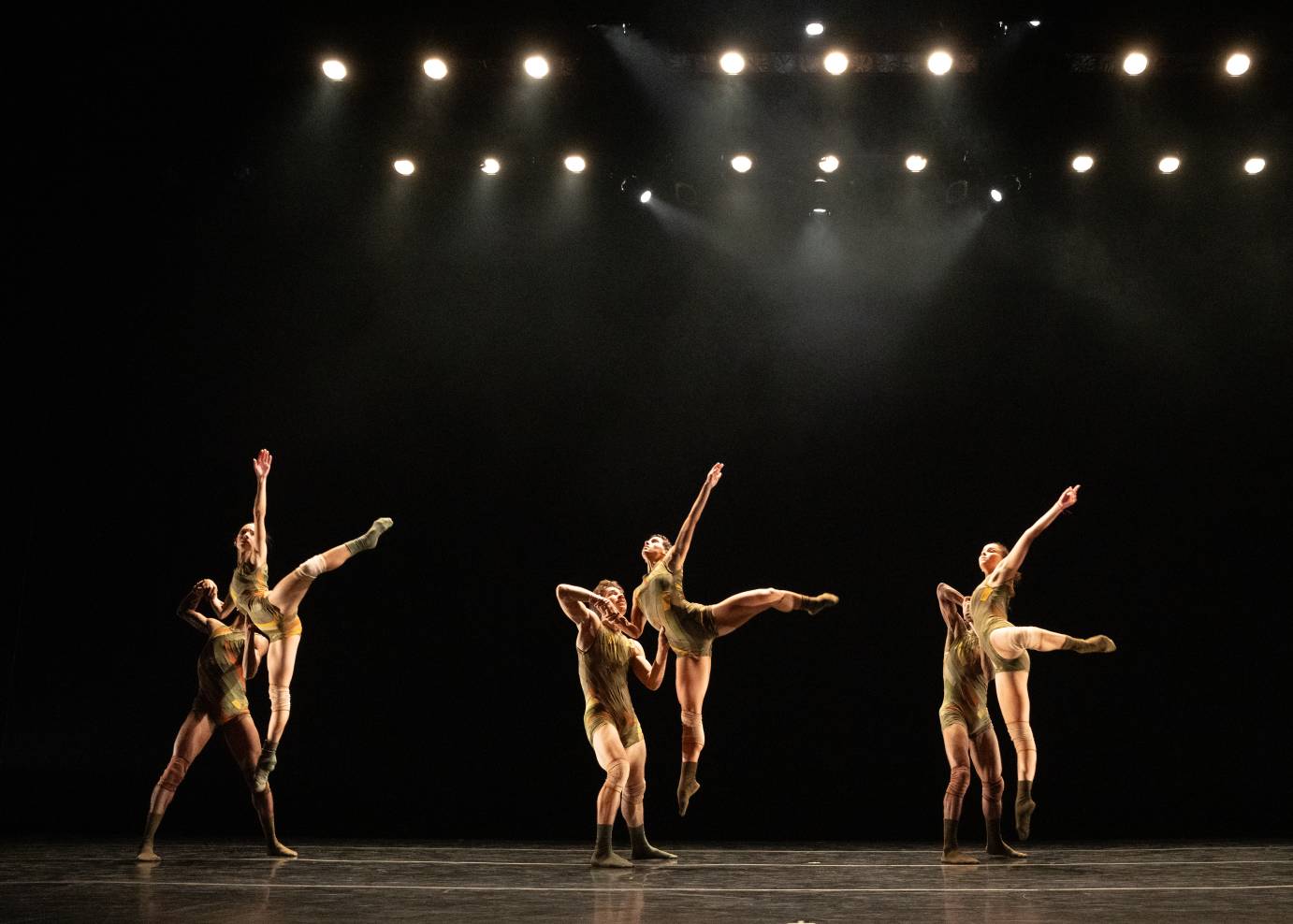Three couples are situated side by side with the front dancer jumping high in the air, one leg extended to the side and one arm thrown skyward
