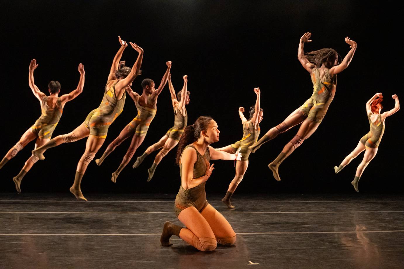 A woman kneels as seven other dancers jump upward, their arms above their heads.