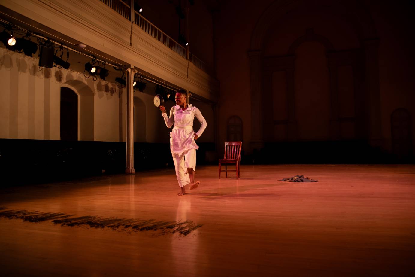 A smiling woman dressed in white, lifting a foot as she walks, holds a frying pan near her face. The space is lined with lavendar sprigs.