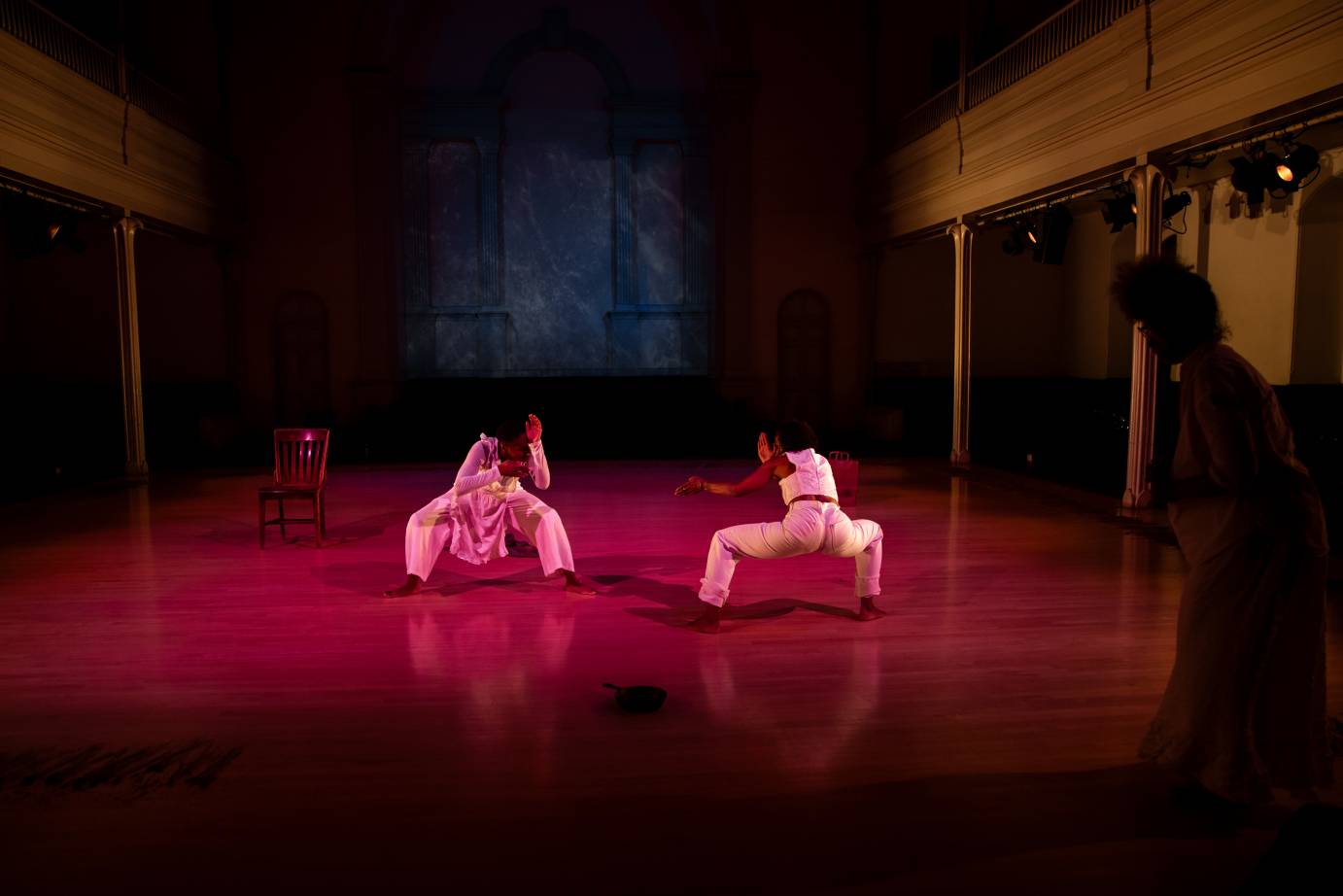 Two women in white, with the brown slatted chair in the background, face one another while crouched in a deep second postion.