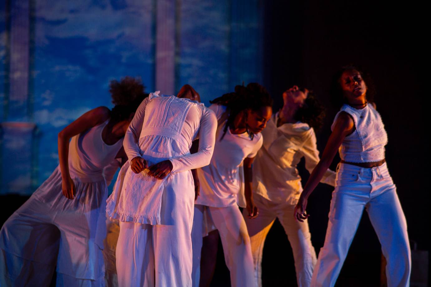  A group of standing performers dressed in white clump together. They look up, out or down.