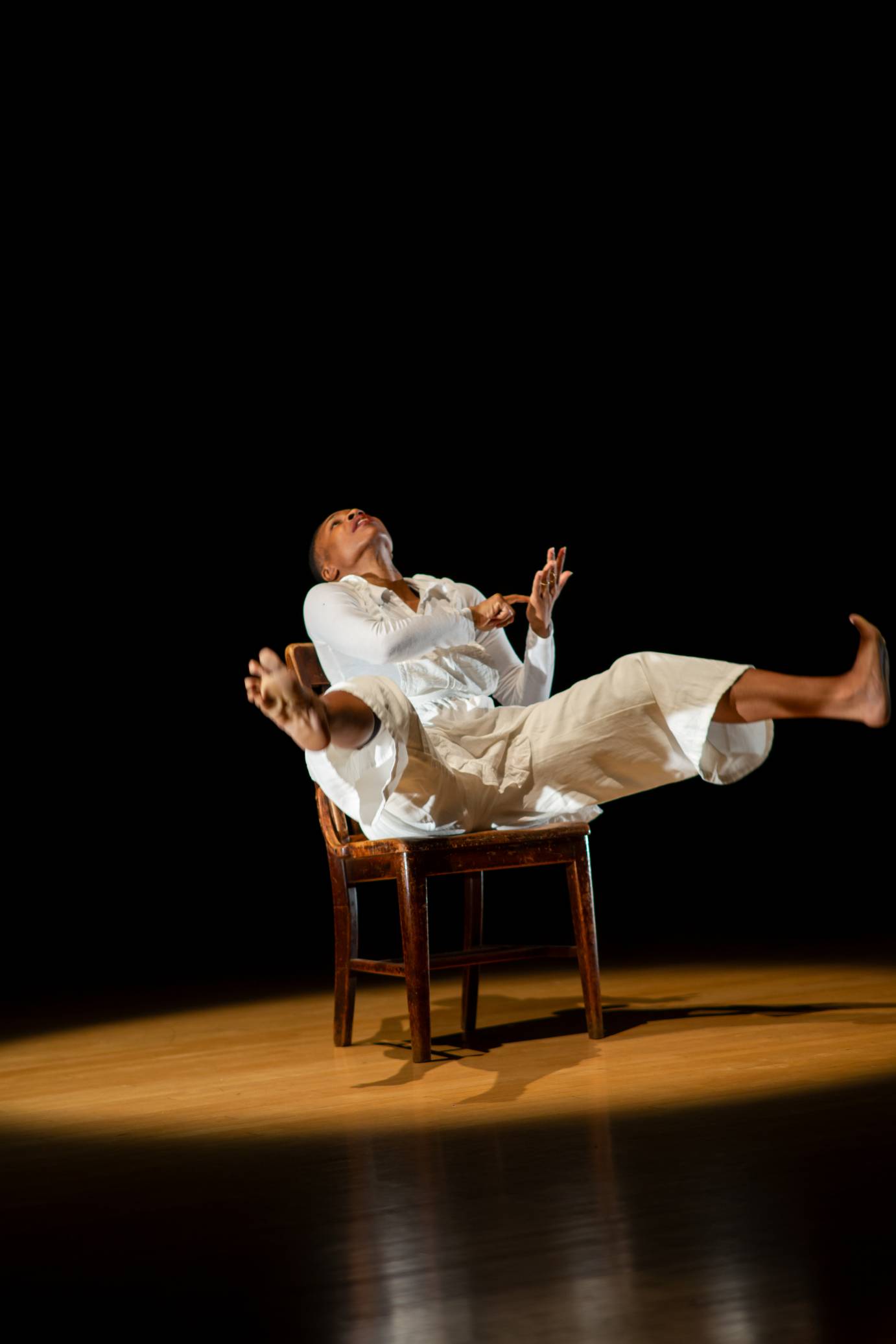 A woman dressed in white, seated in a brown chair, rocks back, legs akimbo, head back, one finger stabbing her other palm.