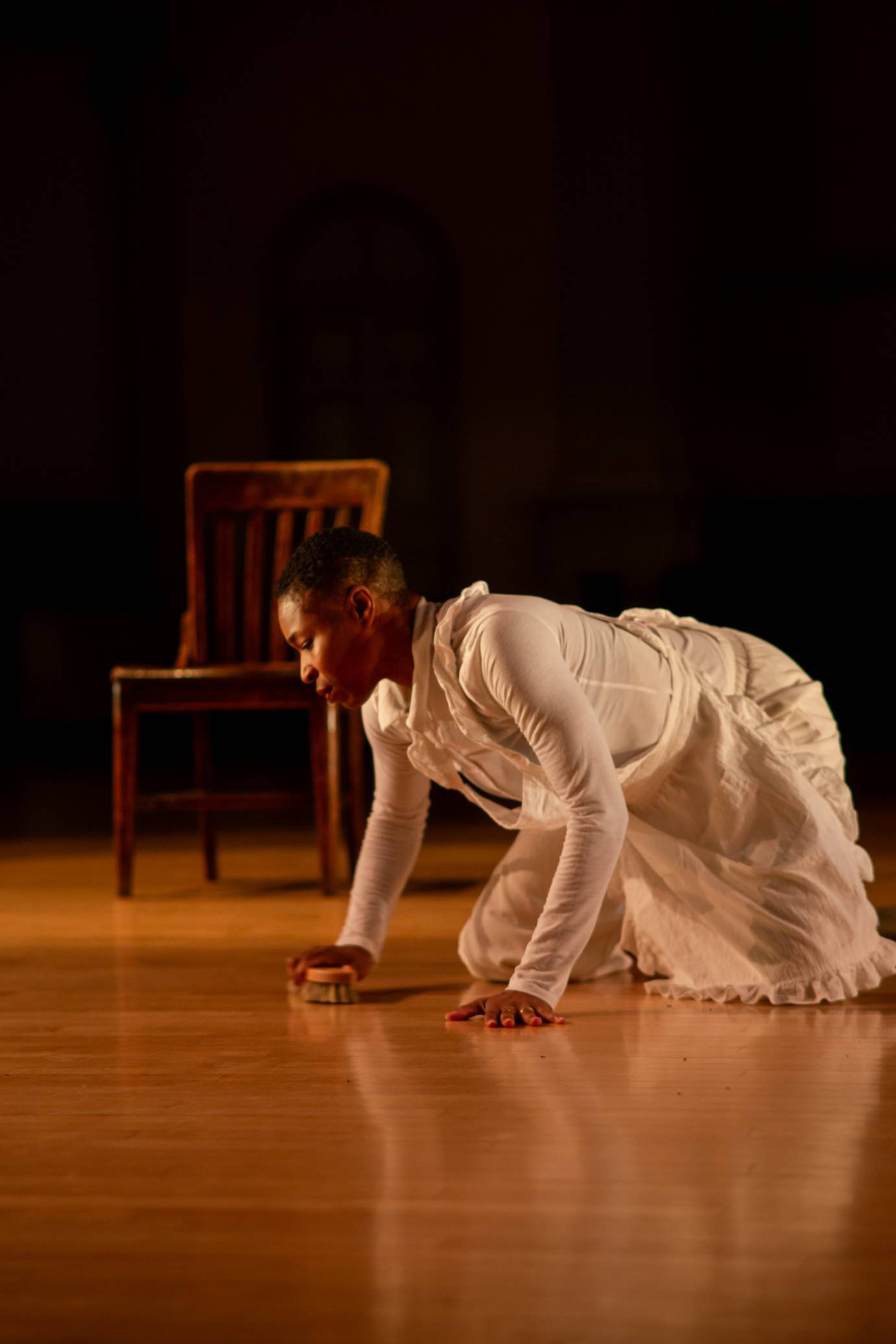 A woman in white, wearing a white ruffled apron, is on the floor on all fours scrubbing. Her slatted brown wooden chair stands in the background.