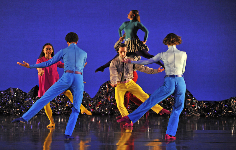 A quart of dancers stand on one leg will the other hovers low to the ground. Another dancer in the background is lifted by a fellow partner..