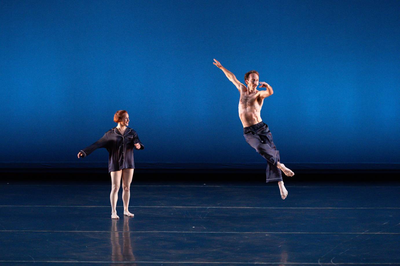 A red-headed woman wearing a long-sleeved navy pajama top smiles as she watches a bare-chested man jump high in a twist wearing the pajama bottoms.