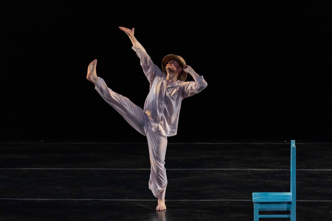 A hatted man dressed in a loose gray long-sleeved top and pants, stands on releve with the other leg kicked upward to match a lengthened arm. A light blue wooden chair accompanies.