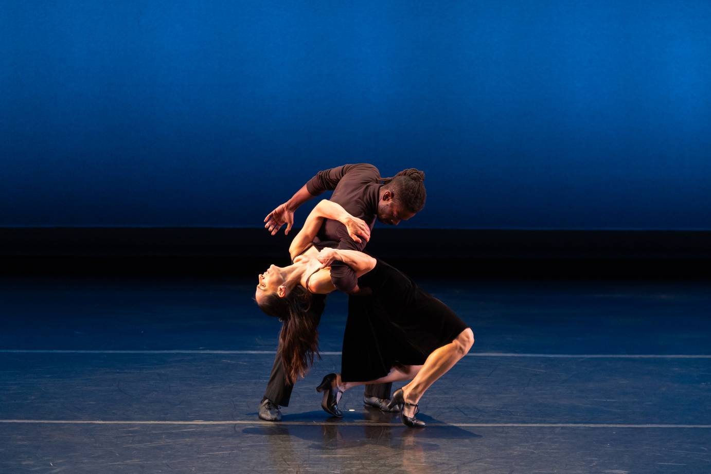 A woman in a sleeveless black dress arches backward held up by a standing man who spirals toward her.