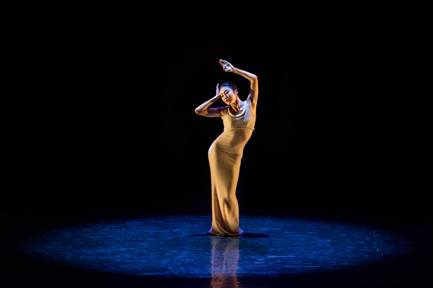 A woman in yellow snugly fit long dress, rests one hand on her forehead and lifts the other in the hair. She stands in a narrow position under a spotlight.