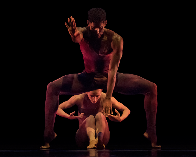 The dark skinned man bends his legs in a large horizontal knee bend, one hand reachers forward as if to say stop the other arm and hand reaches down between his legs. Beihind him is a light skinned woman seated.