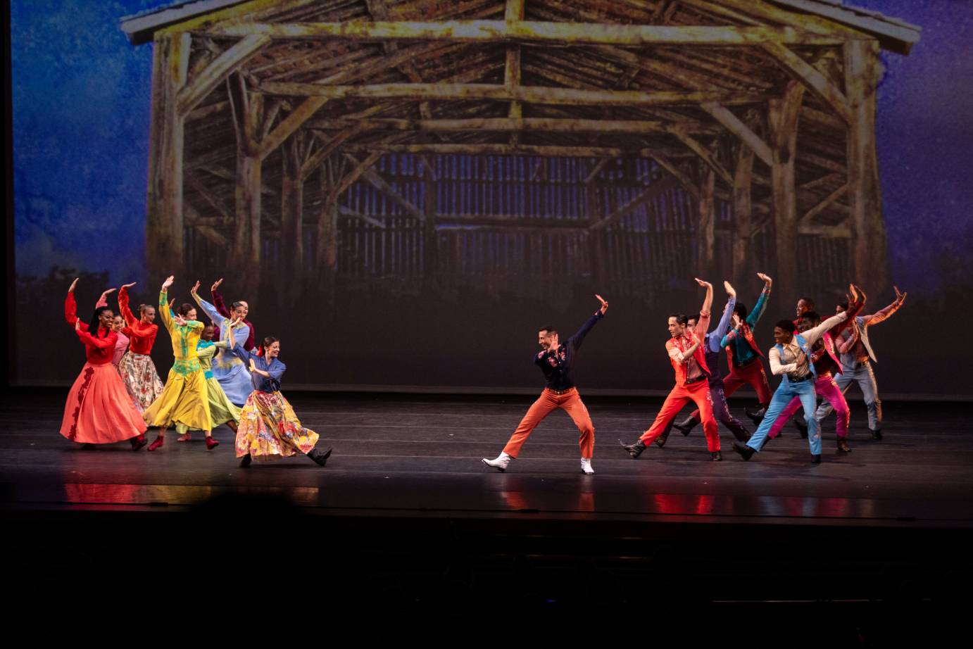 The company in colorful long dresses for the women and pants and shirts for the men, dance in separate gendered groups in front of a projection of the inside of a barn structure. A woman and man in unison with the two groups dance in front.