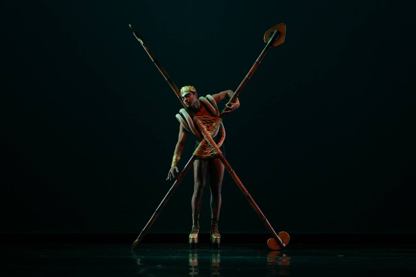 A man wearing a crown, standing in his gold platform shoes, leans on two crossed sticks.