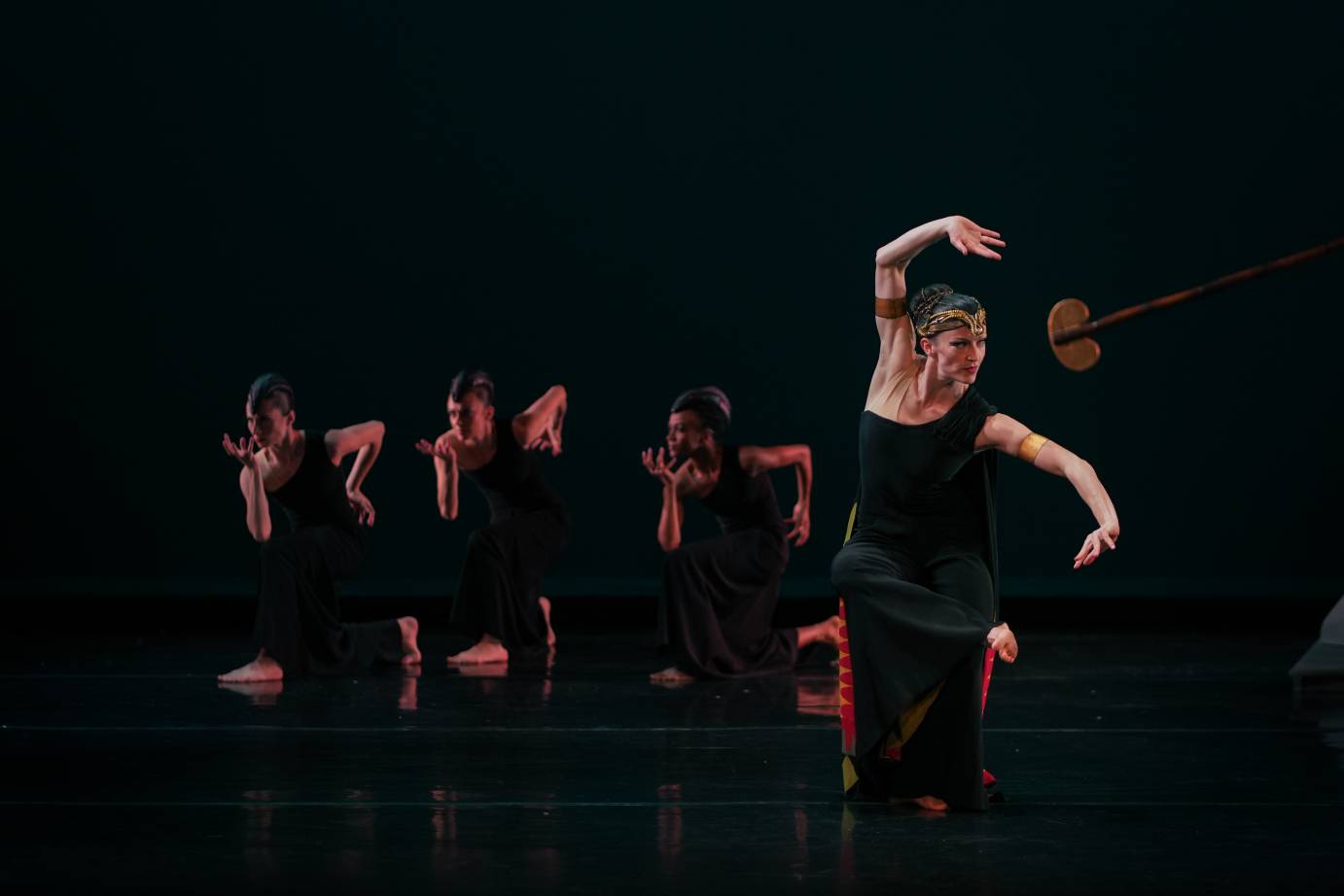 A woman with an intense focus to her left, arm bent overhead and kneeling in a black dress, is backed up by three upstage dancers whose bodies are facing the opposite direction. Their bodies, also dressed in black, are bent forward, hands below chins and near their hips.forward
