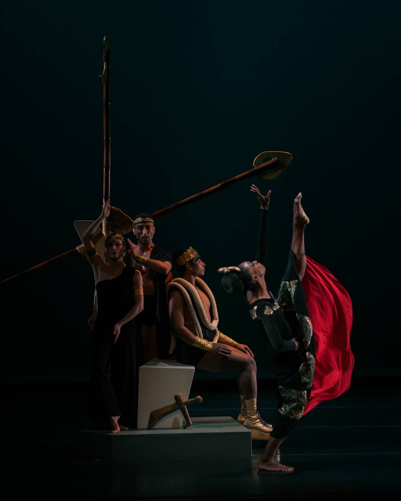 An elaborate set piece with the man in platform shoes seated on a stylized throne while a woman in a red cloak stands on one leg and lengthens her other fully vertical to the ceiling. Two figures. behind the king, look to the audience encumbered by the sticks. A sword rests against the throne.