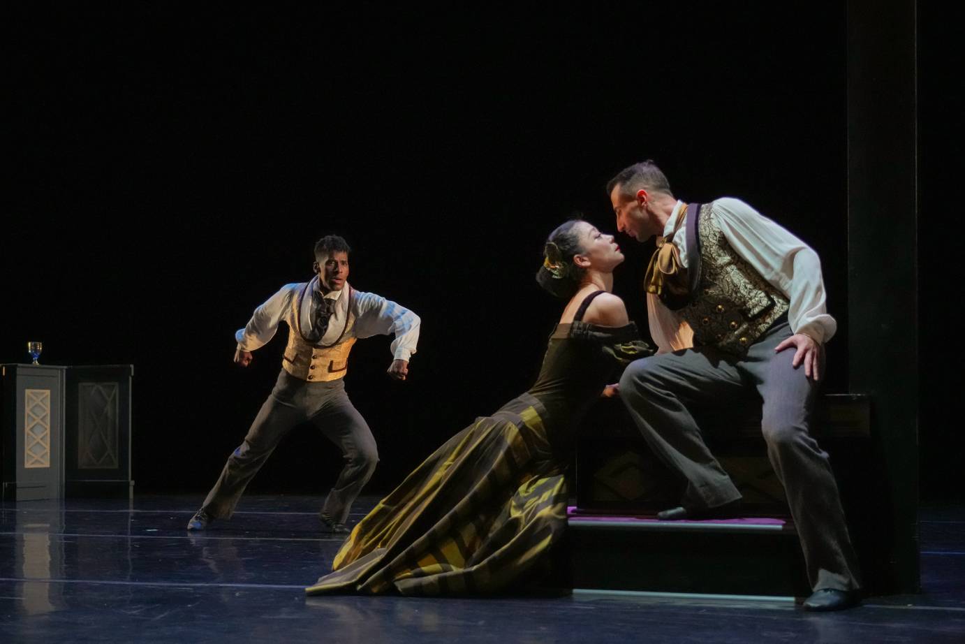 The faces of a woman and man are close together while a third man looks on in confusion. All are dressed in costumes from the 19th century - the men wearing vests and white shirts with billowy arms, the woman in a full dress off the shoulders.