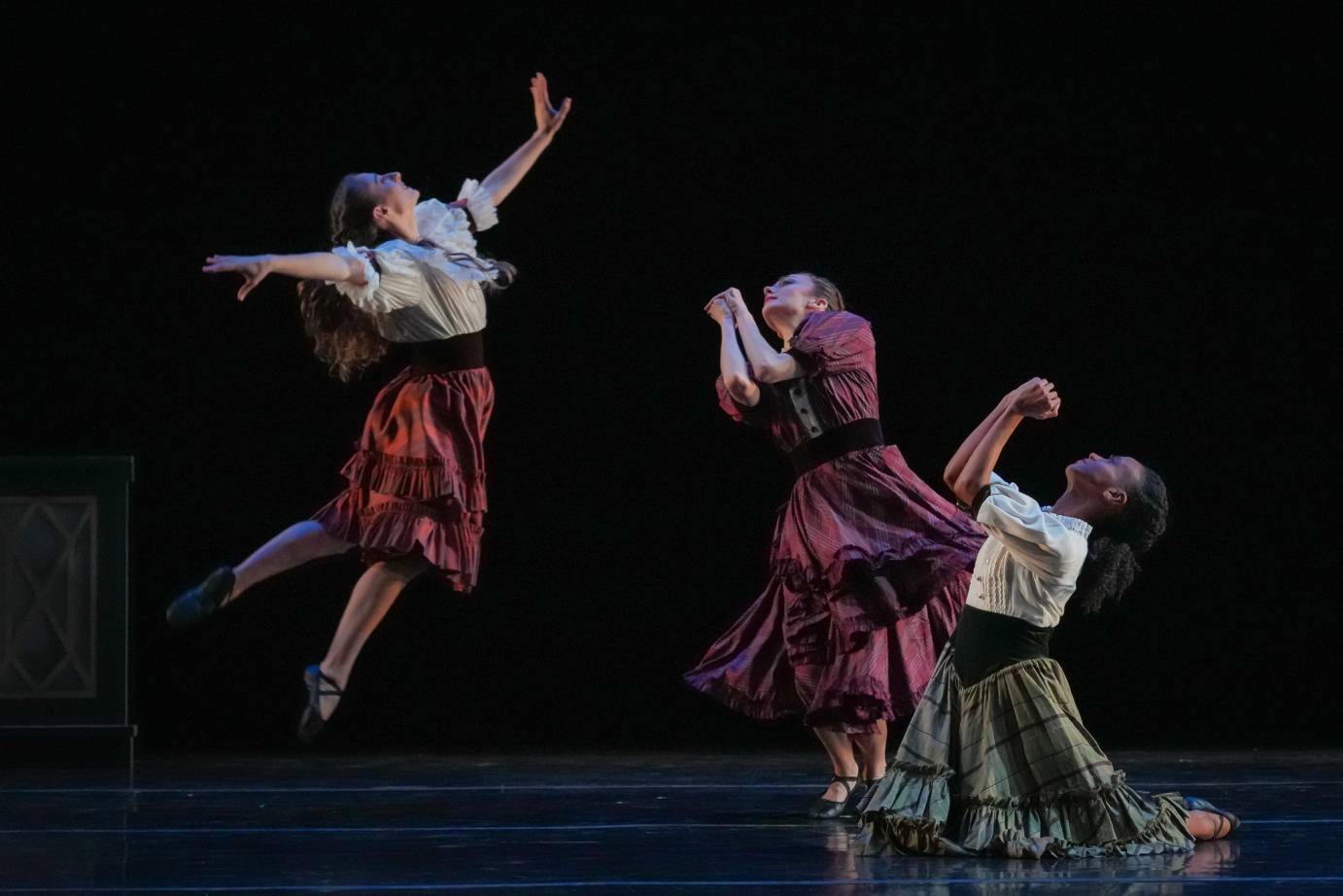 Three sisters, dressed in flowing skirts and blouses, are in various stages of release. One sister kneels with hands clasped as if praying. A second sister stands and spirals with elbows bent and together while the third sister has jumped off the ground. All three faces are tilted upward.