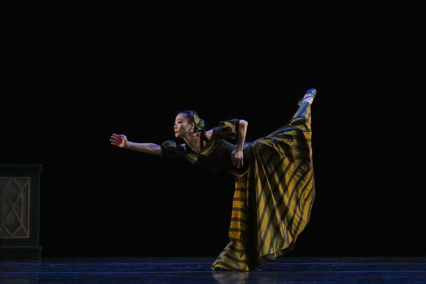 A woman standing in arabesque thrusts her body far forward. One arm is extended while the other is bent. Her leg behind is high and long.