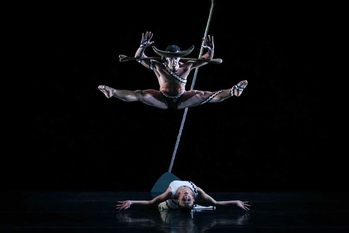 A man in a half-moon like headdress with legs outstretched in a V hovers above a woman lying on her back with her head turned upside down to the audience.