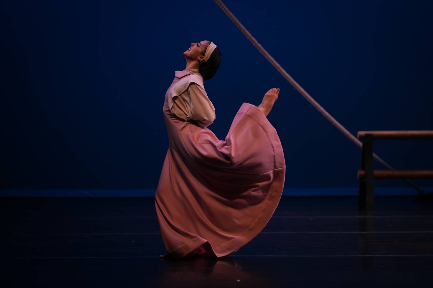A woman dressed in a long, 'old-fashioned' pink-lit dress arches toward her foot that reaches toward her head. Her facial expression is ecstatic. A rope line and wooden set are close to her body.