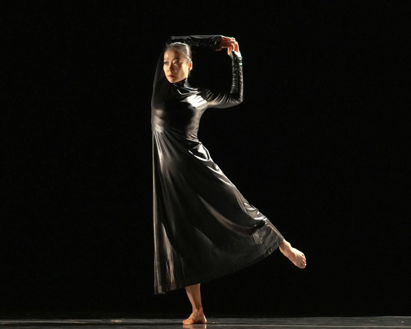 Woman standing on right let with right arm draped over head, hands clasped. With a determined expression, the dancer is wearing a silvery black turtle neck costume.