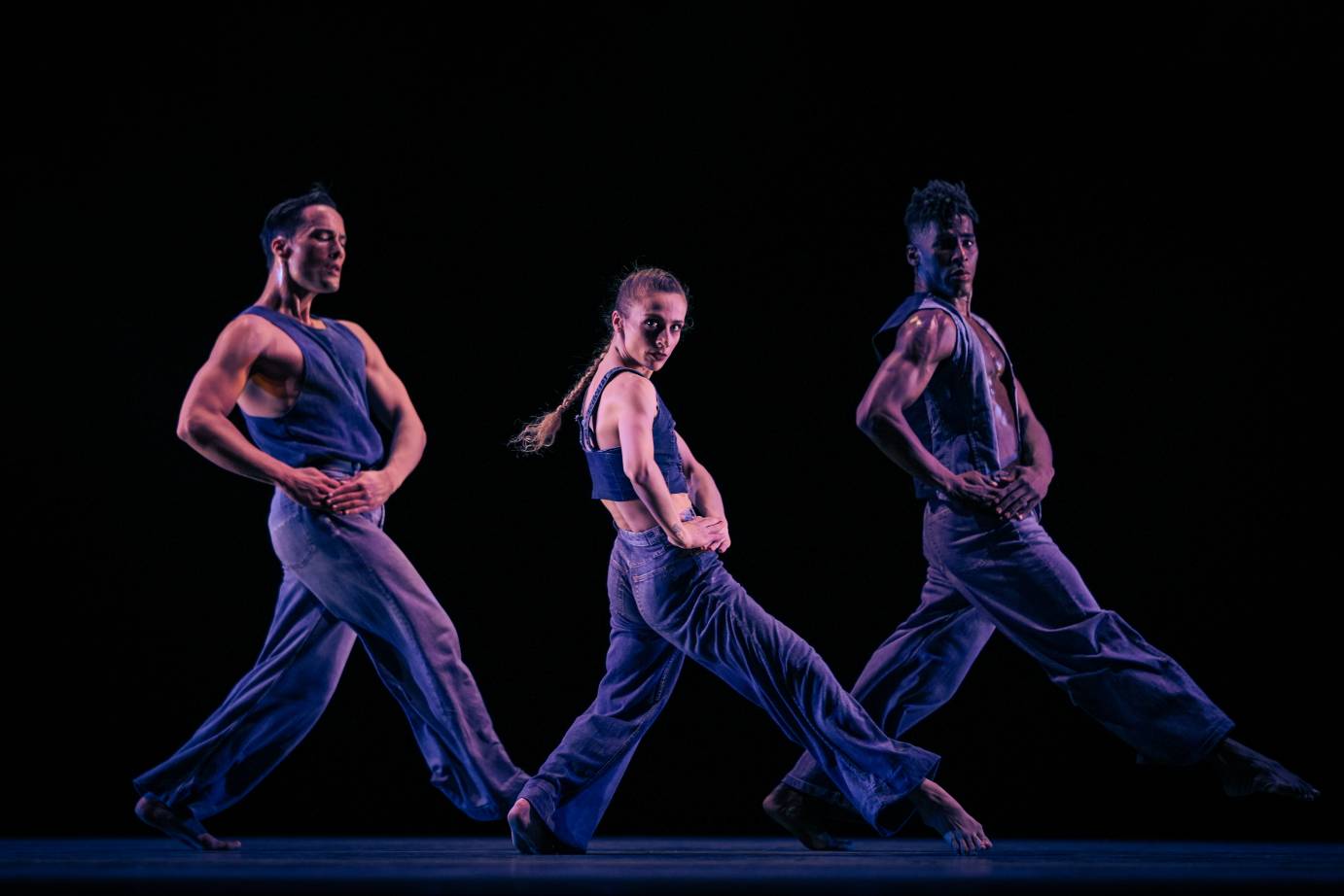 Three dancers, one woman between two men, in unison facing stage left, looking out to the audience with both hands on right hip. Costumed in blue pants and sleeveless tops.