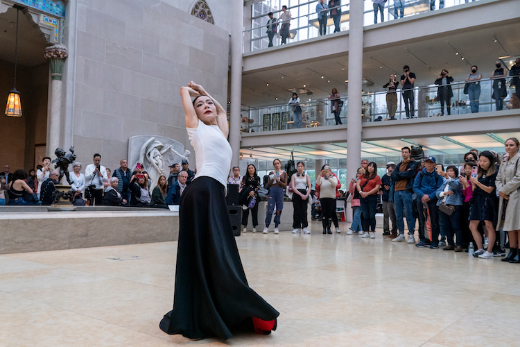 woman with tight black bun arms framing the sides of her face, in a stage of deep concentration and purposefulness as the audience looks on