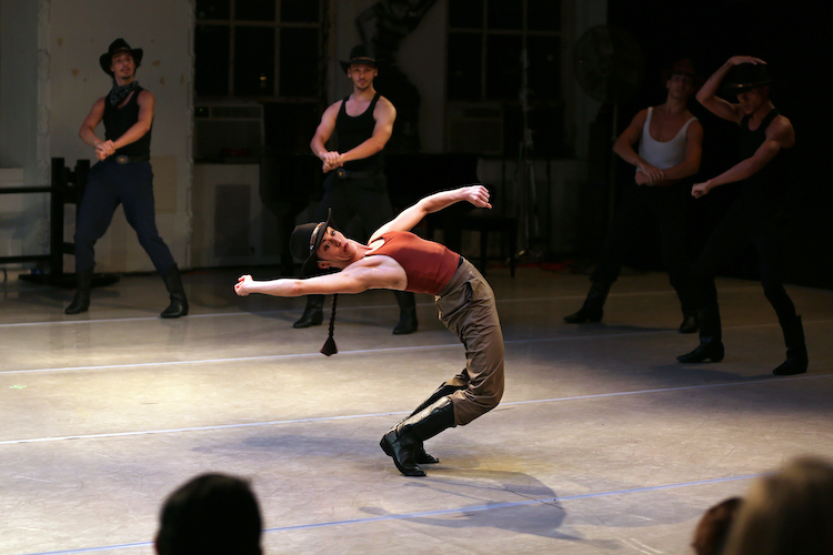 the cowgirl character of Rodeo, is center stage and expressing her awkwardness as she stands on her toes and leans backward with an incredulous expression on her face 