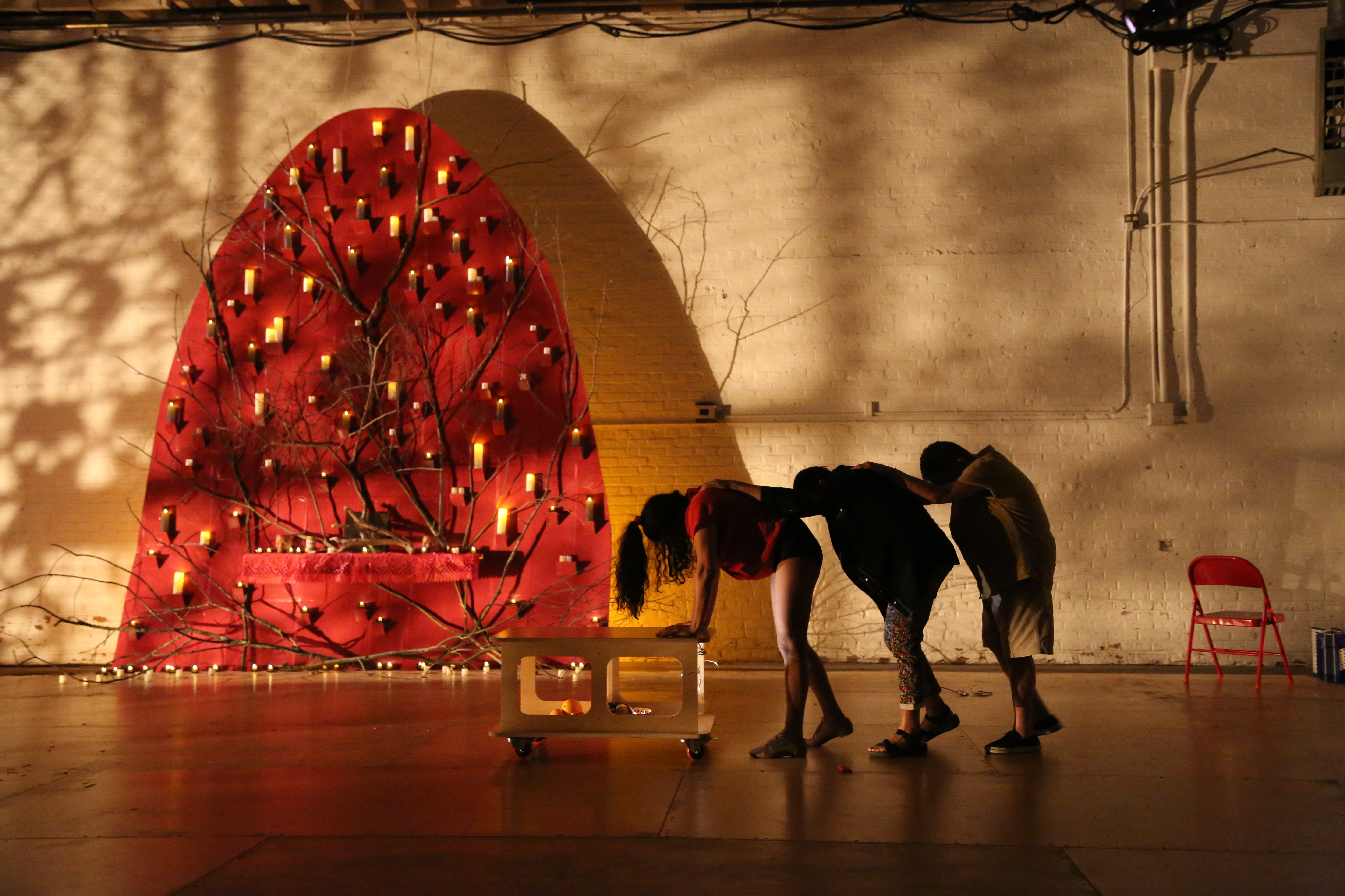 Three figures in silhouette place arms on the person's shoulders in front of them. The lead person leans on a short table. A red shape situated upstage acts as a set.