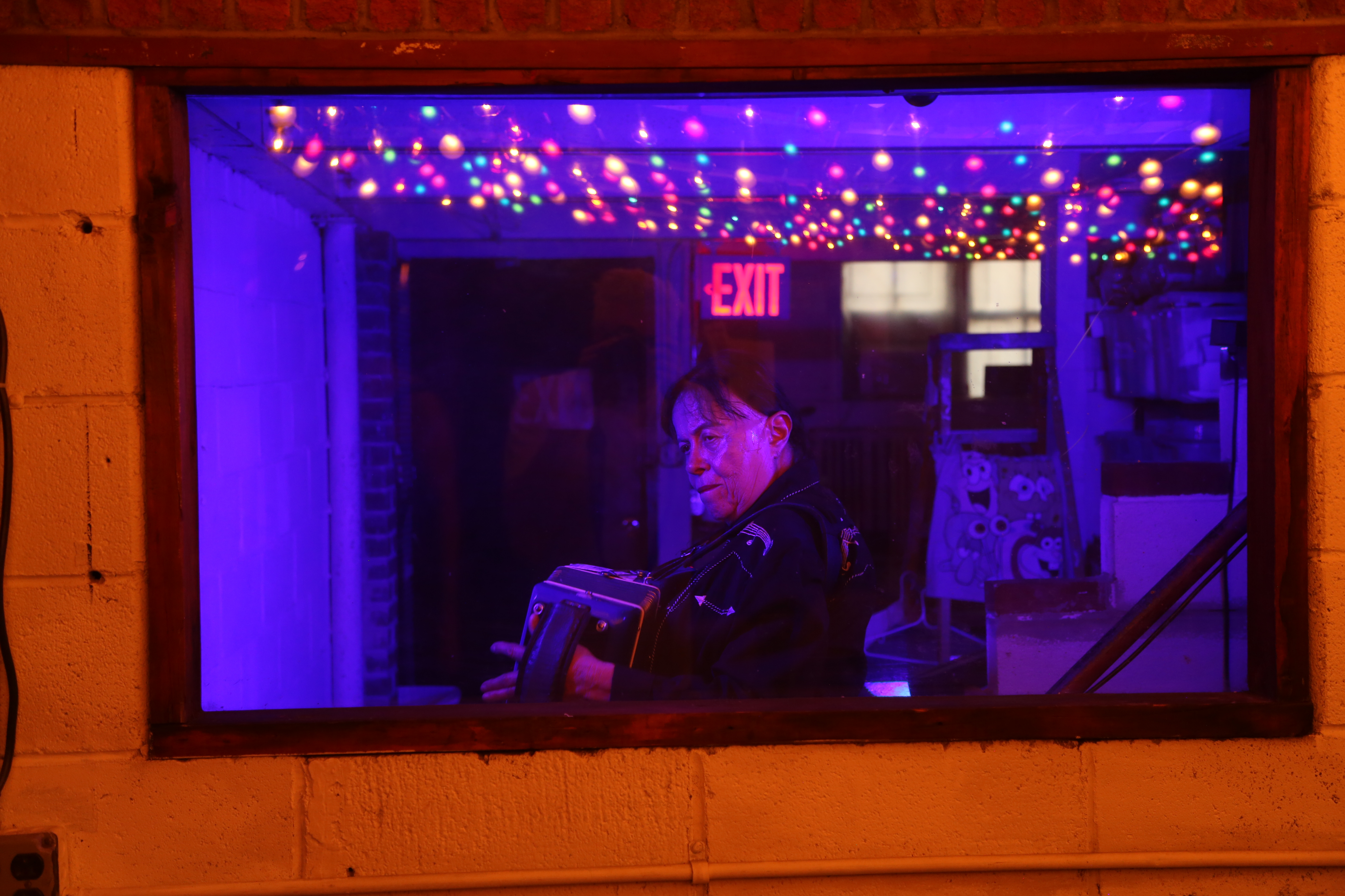 A man, inside a room with an exit sign and small colored lights hanging from the ceiling, plays an accordian.