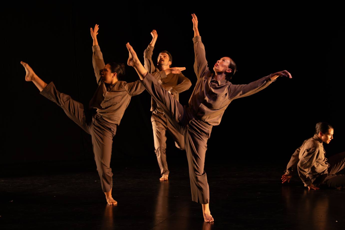 Three dancers close together: two women and either side of a man wearing dun colored shirts and pants, toss arms skyward with extended legs.