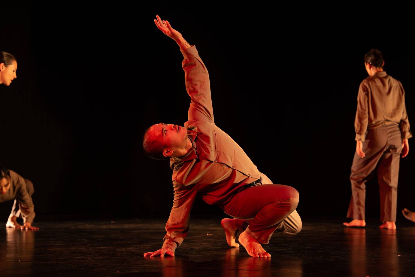 A mustachiod man, in long-sleeved dun colored shirt and pants, squats reaching one arm long. His gaze following. 