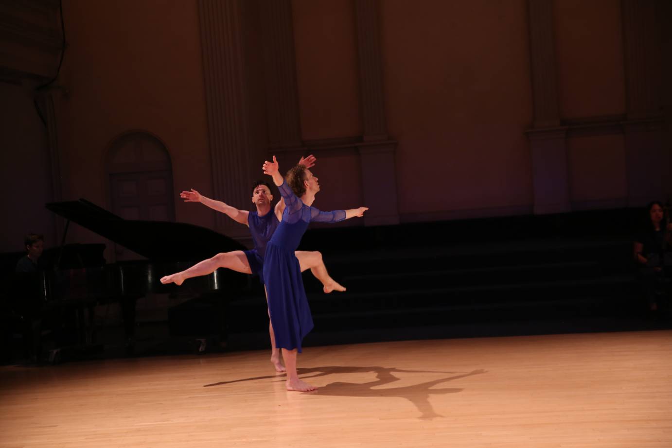 Two dancers on releve, close together, each lift one leg on attidude while both arms reach.
