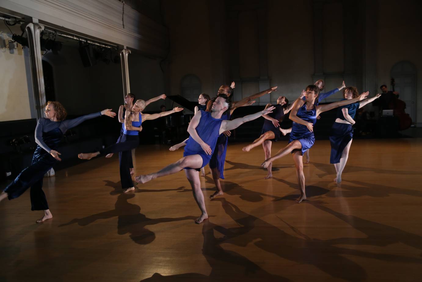 A cast of eleven dancers dressed in blue costumes of various shapes, stand on their right legs on the diagonal close together. Their left leg is lifted in front attitude while their left arm is straight on the diagonal. The right arm is wrapped around their torsos.