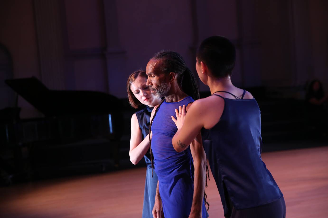 Two women stand on either side of a man. His eyes are lowered and his face is contemplative. The women place their palms on the sides of his body.