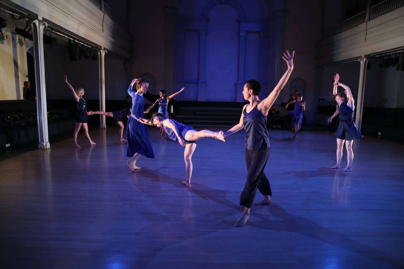 Eight dancers connected and disconnected - one in arabesque and others standing with their arms stretched high - are bathed in blue light.