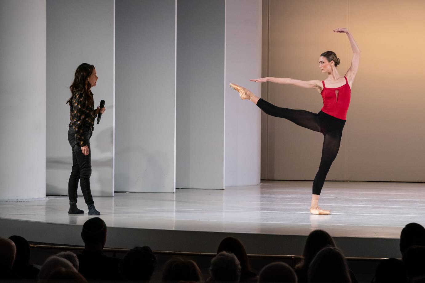 The choreographer holding a microphone works with a woman dancer on pointe with her right leg extended into second as she stands on a flat pointe shoe.