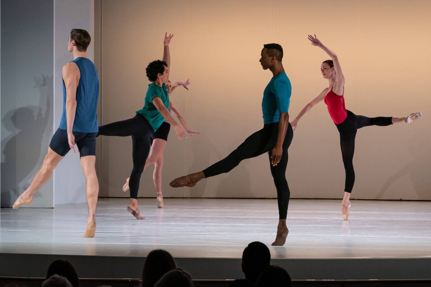 Five dancers dressed in black bottoms and red, blue and green tops, loosely face in different directions while on releve either in pointe shoes or ballet slippers. Two women face one another while two men look in the direction they are traveling.