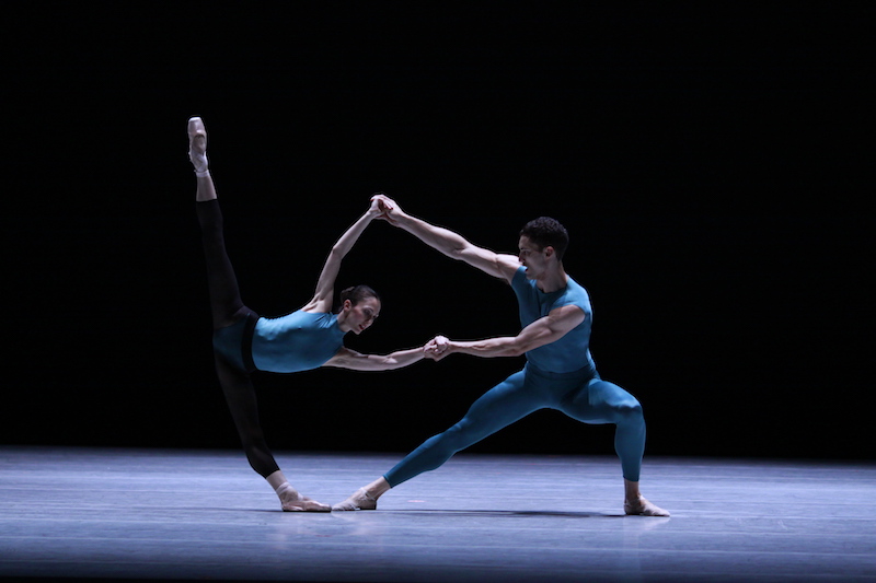 A couple clasp one another hands. Both leaning away from the center, the woman développés her right leg to a 180 degree angle. 