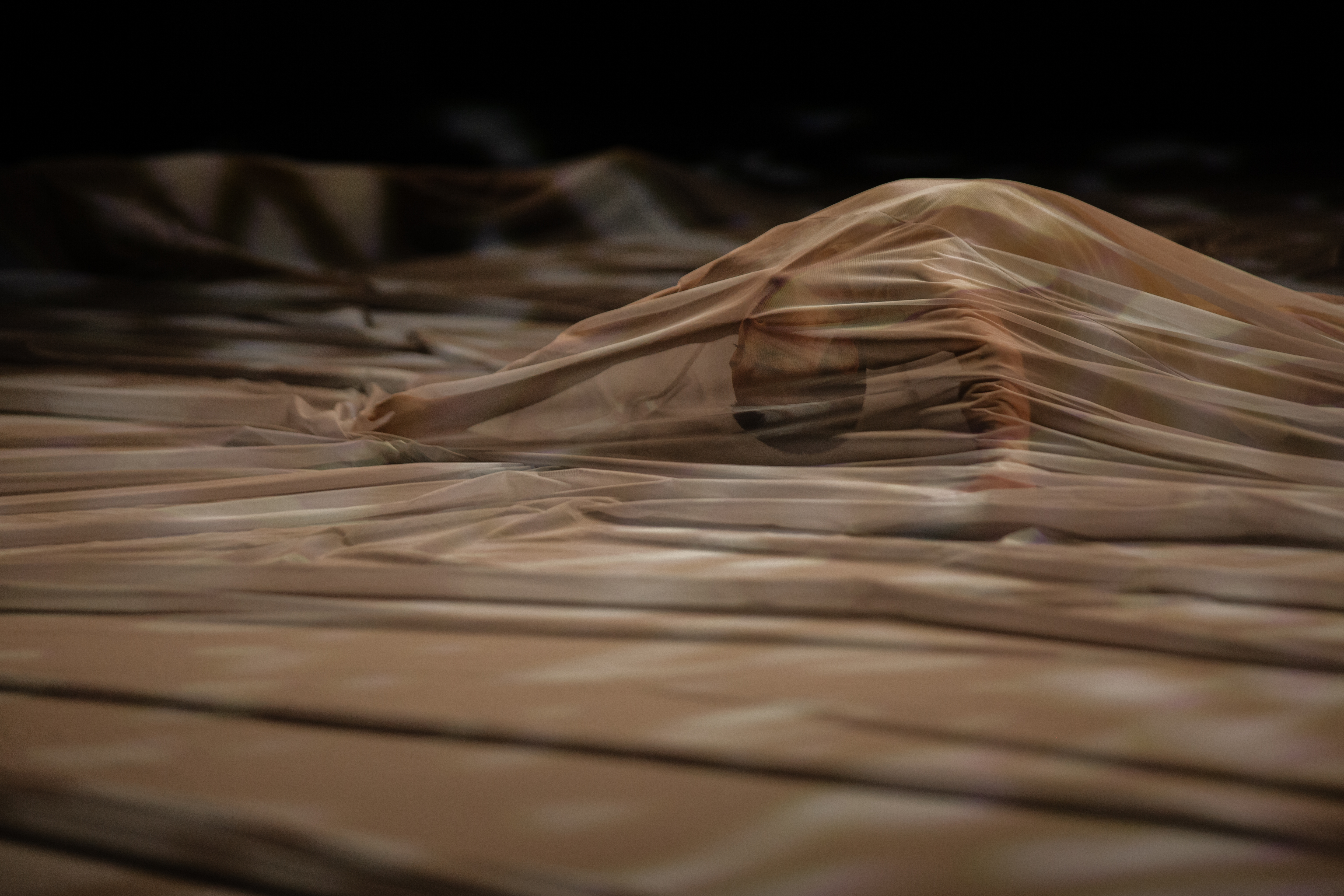 A performer is seemingly upside down on the stage floor beneath a tan-colored, dappled, opaque fabric