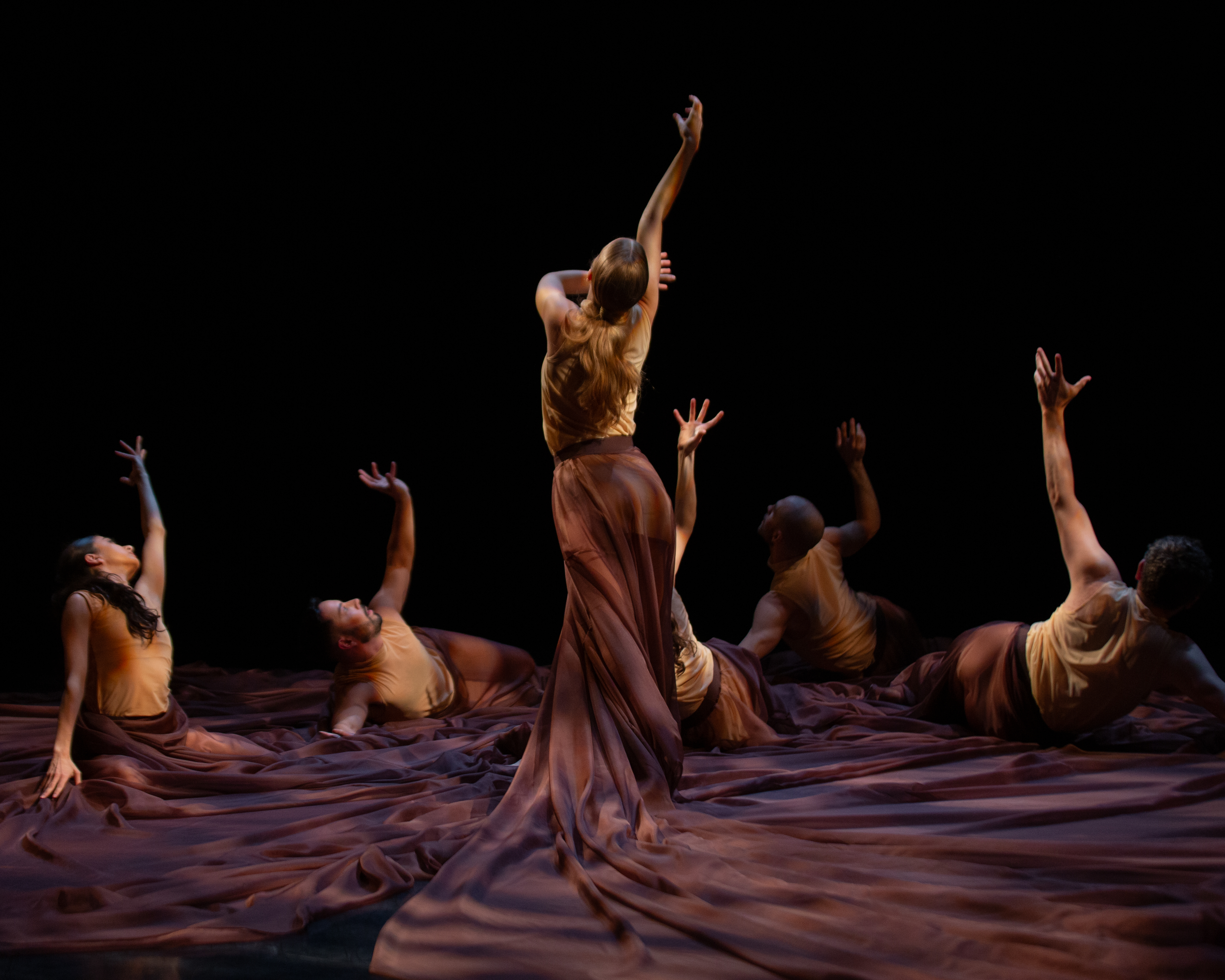 One standing dancer with long, auburn hair is surrounded by five others spiraled on the floor, lying on their sides or kneeling, enveloped in purple fabric that entwines the group.